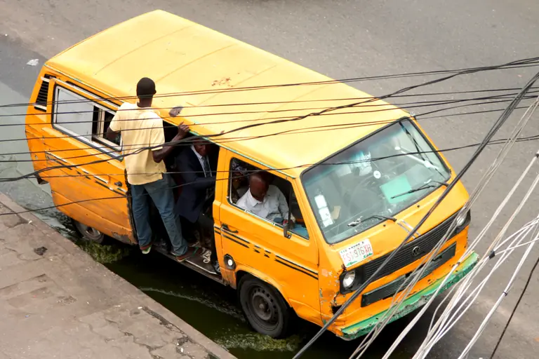 Seen from above, a conductor stands in the open door of a yellow Lagos danfo minibus while the driver waits at the wheel.